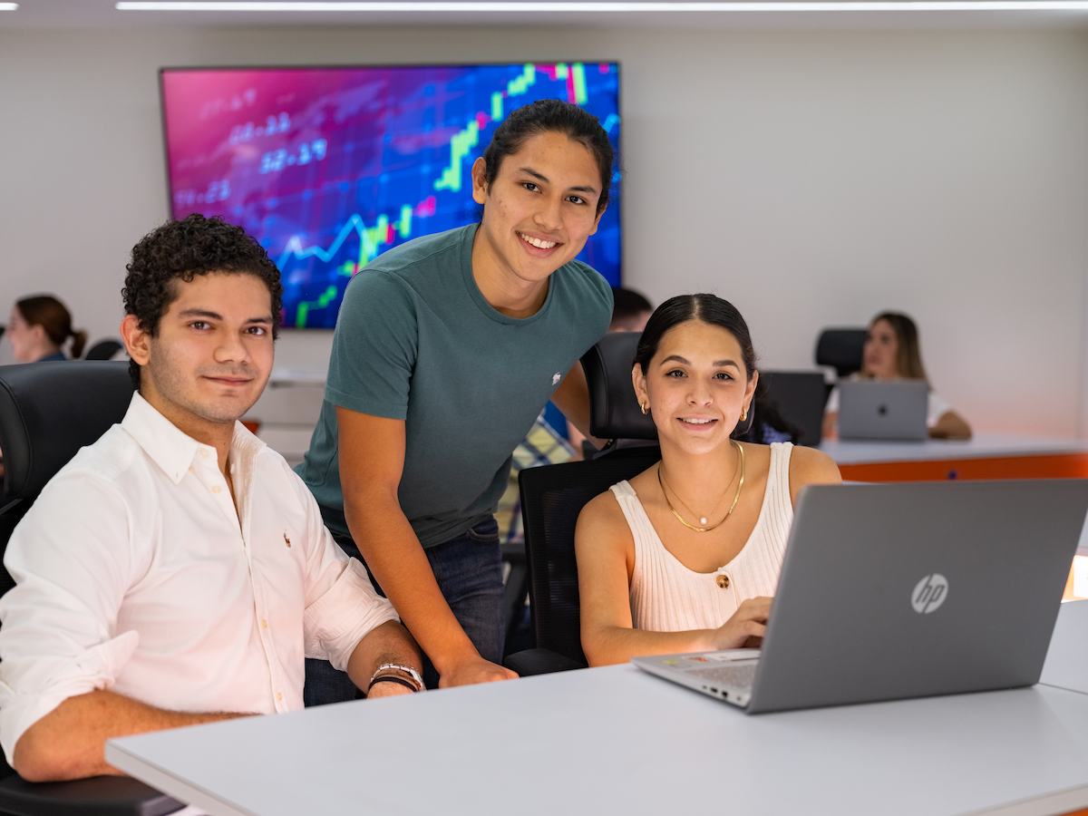 Tres estudiantes posando juntos frente a una laptop en un aula moderna, con gráficas financieras en una pantalla al fondo.